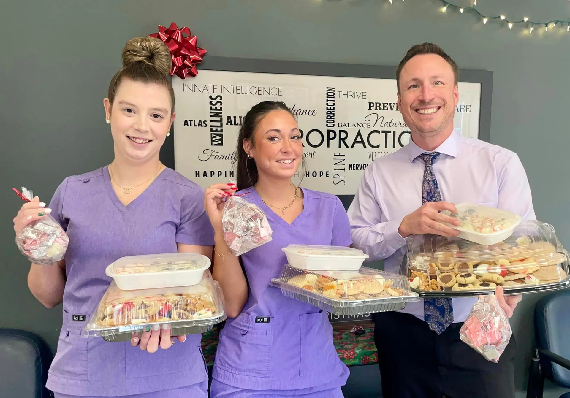 Chiropractor and two team members in purple scrubs holding trays of holiday treats