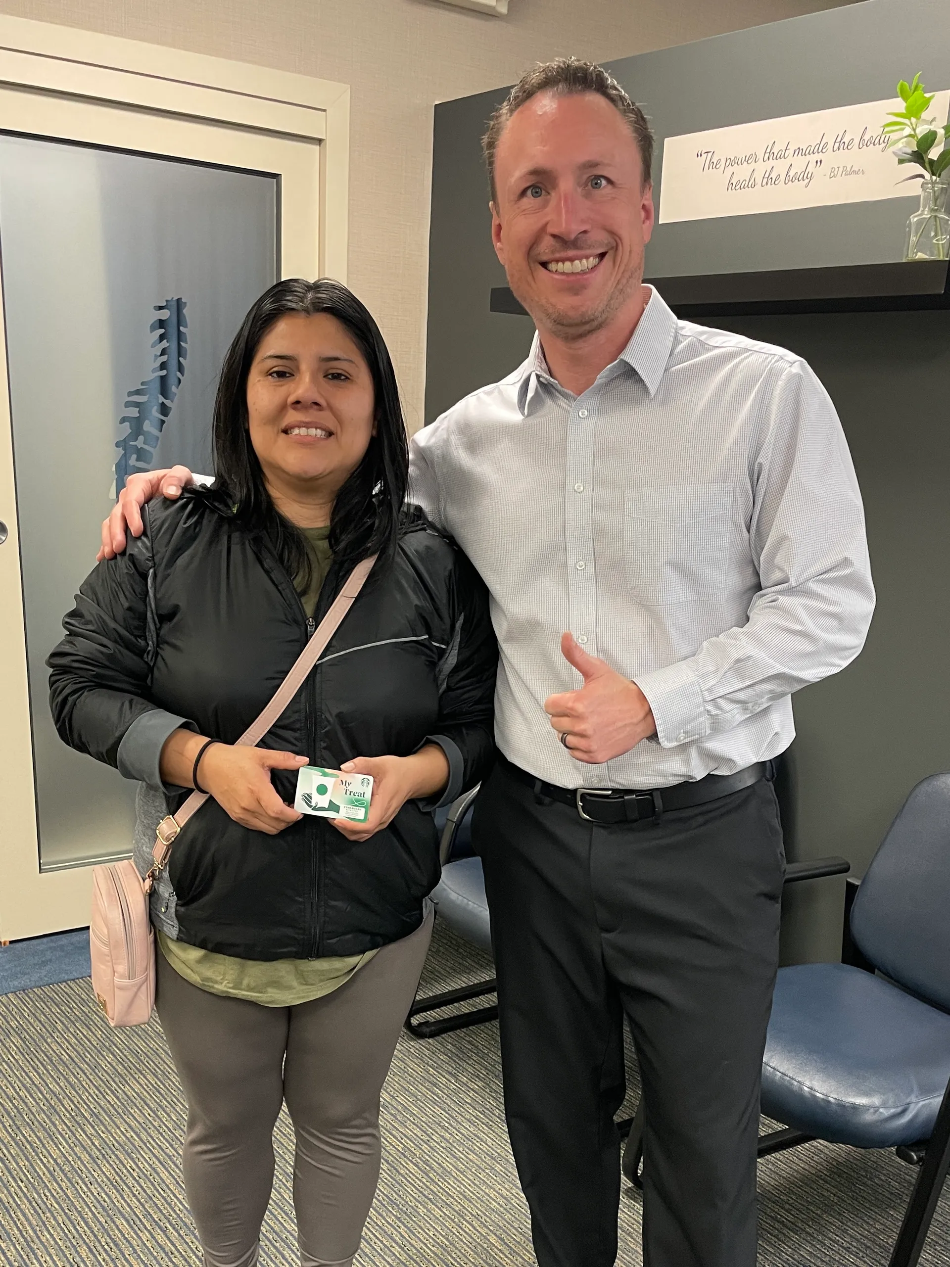 Chiropractor giving a thumbs-up beside a smiling patient holding a card