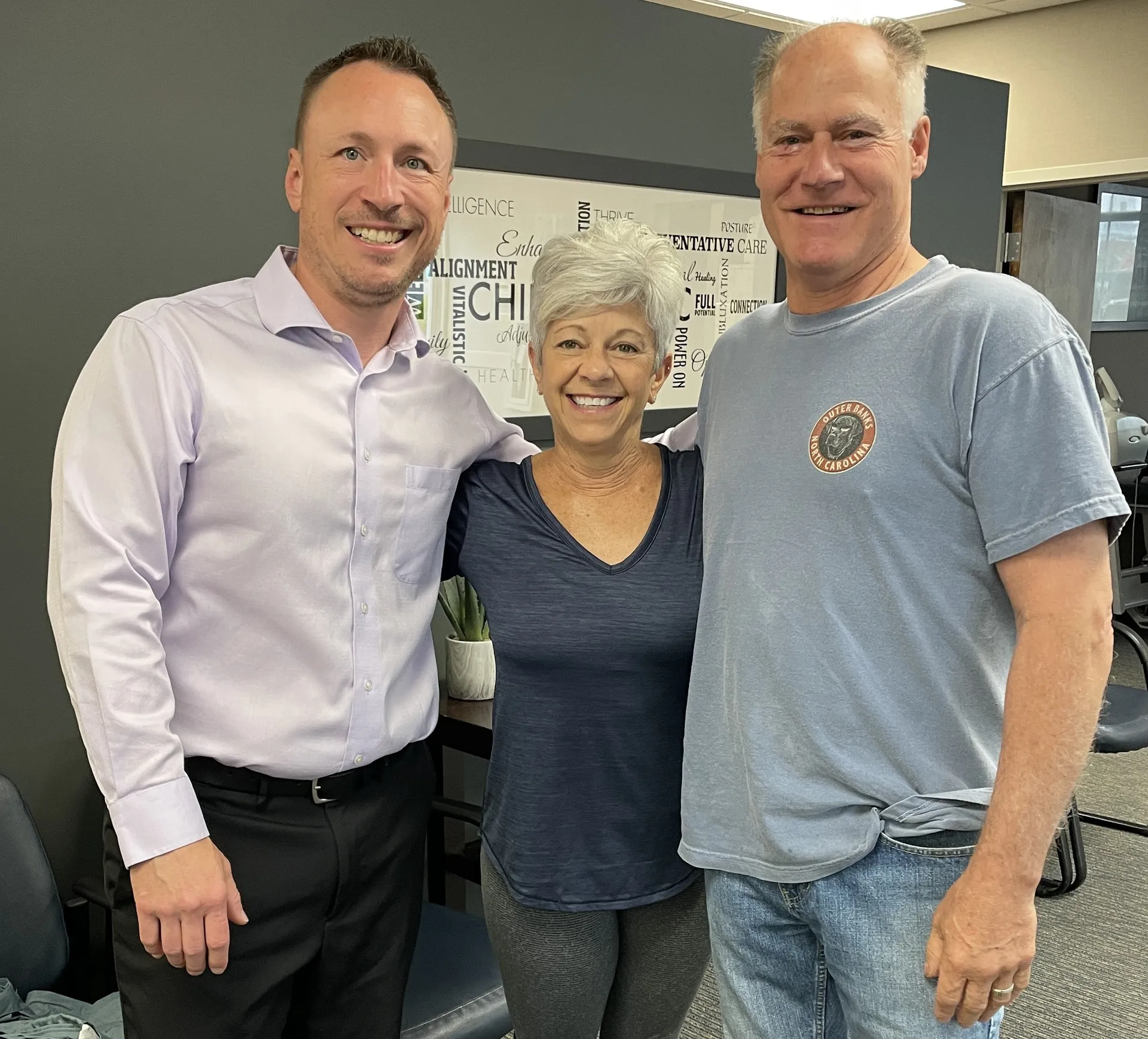 Chiropractor in a lavender dress shirt standing with two smiling patients in the clinic