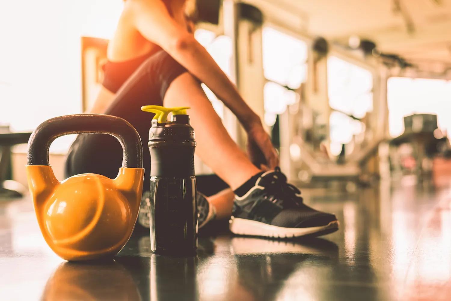 Kettlebell and water bottle on a gym floor beside a woman tying her shoe
