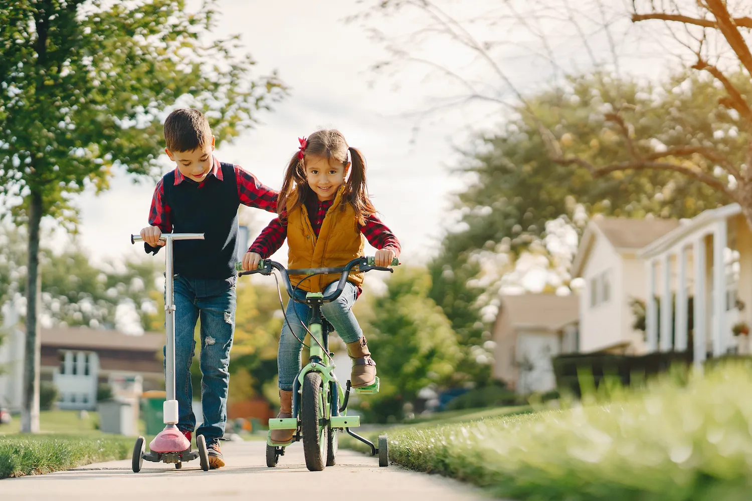 Boy on a scooter and girl on a bike with training wheels riding down a sidewalk