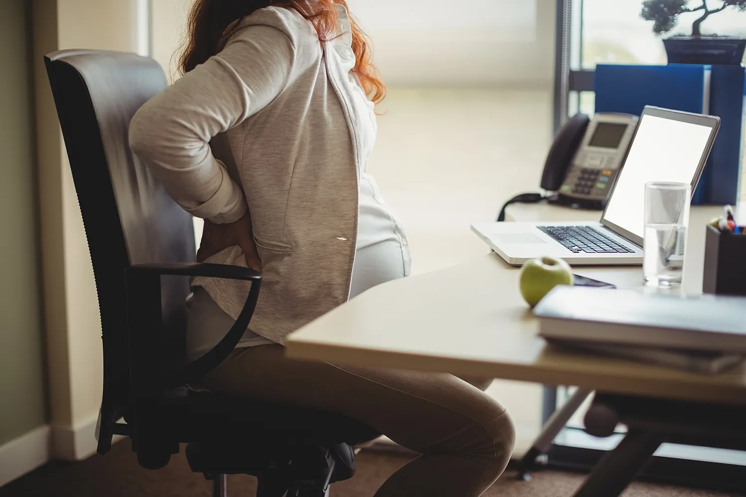 Pregnant woman in an office chair at her desk holding her lower back