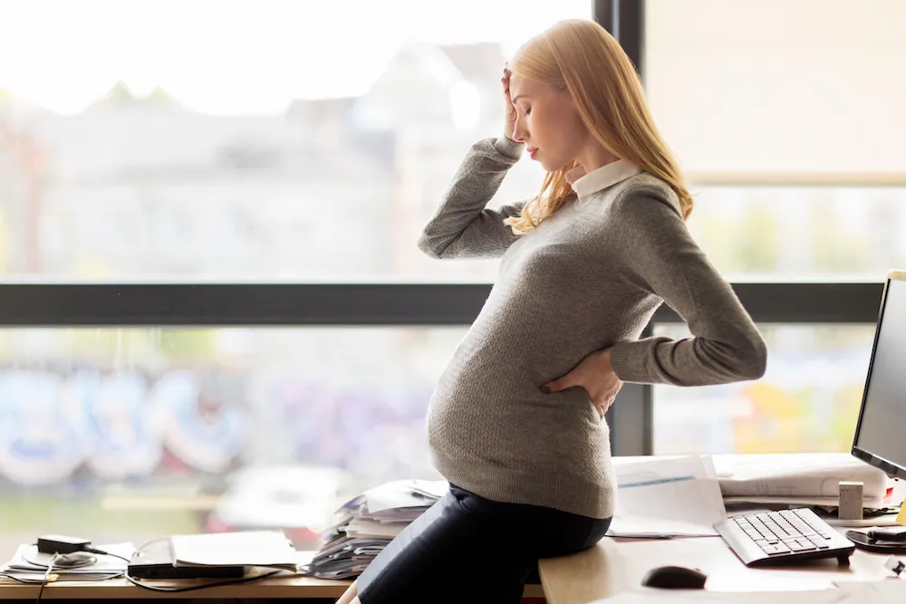 Pregnant woman at her desk holding her lower back and forehead
