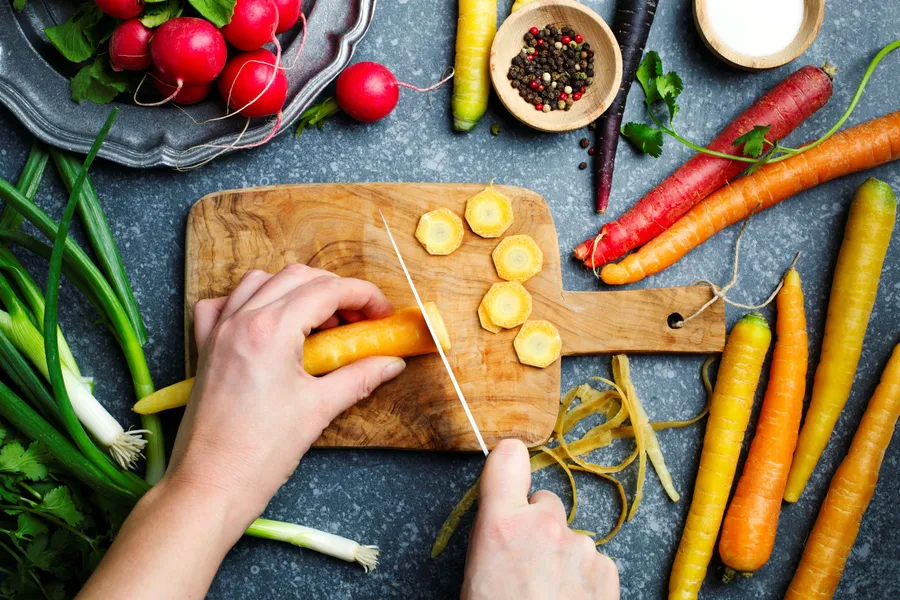 Hands slicing carrots on a cutting board surrounded by fresh vegetables