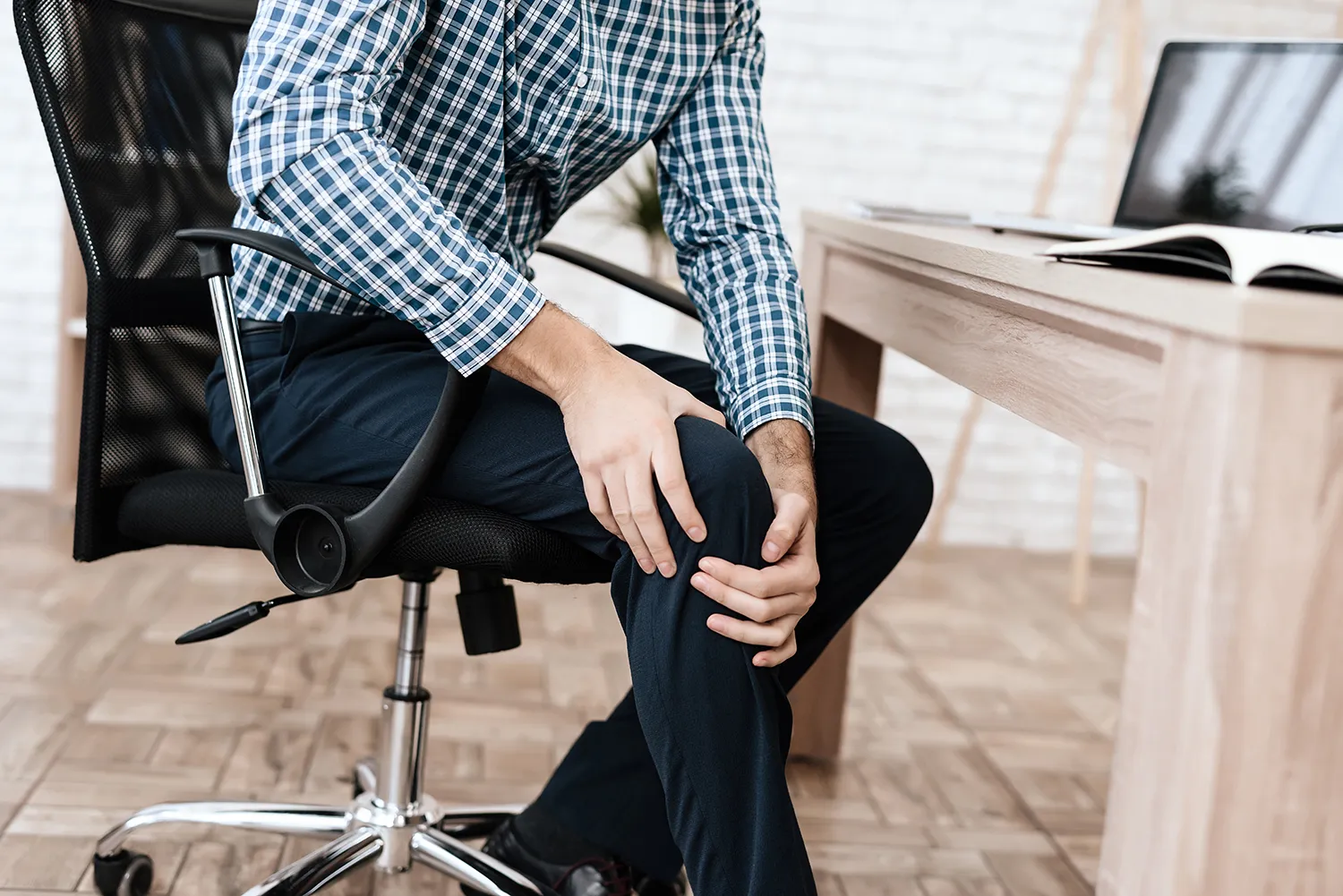 Man in a plaid shirt seated in an office chair holding his knee