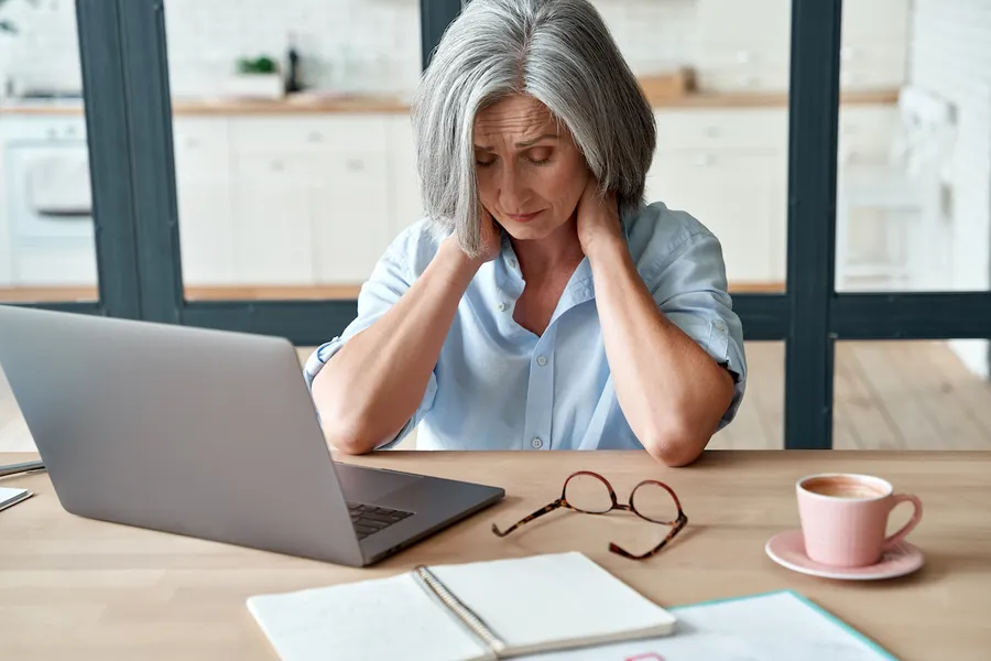 Stressed woman at a laptop holding her head and neck with her eyes closed